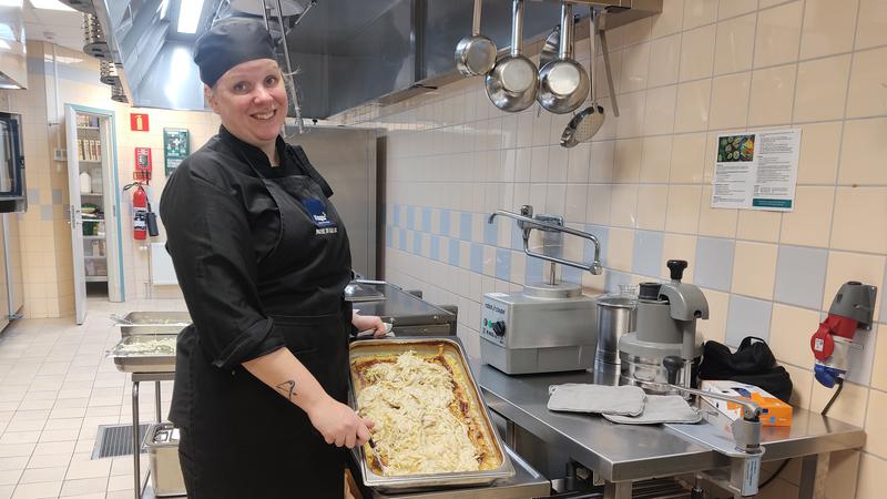 En kock står i ett storkök och håller upp en bytta med mat.