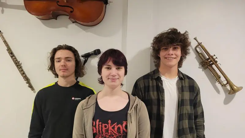 Tre unga personer står framför en vägg där det händer olika musikinstrument.