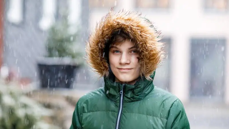 Ung kille i grön jacka med luva står utomhus där det snöar.