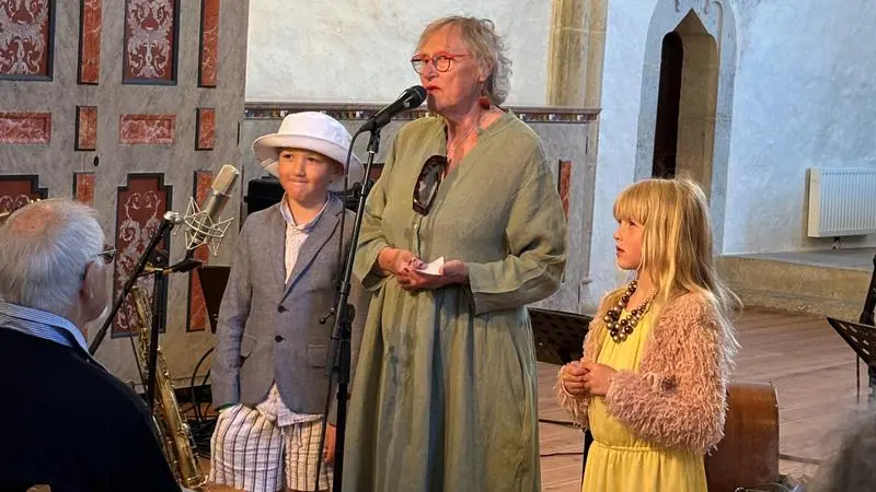 En kvinna med ett barn på varje sida står på en scen i en kyrka framför mikrofoner.
