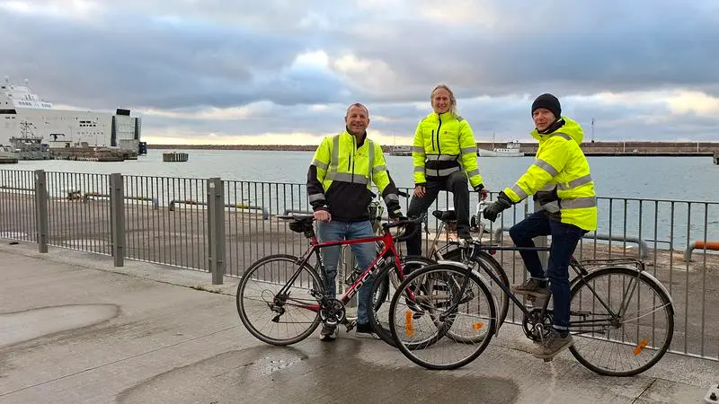 Tre personer i gula arbetsjackor på sina cyklar intill en hamn.