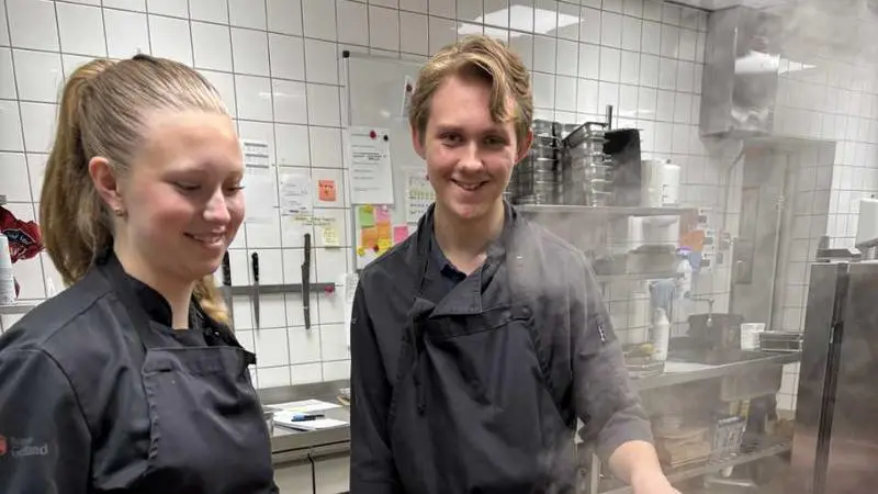 Två unga personer steker lök och morot i storkök.