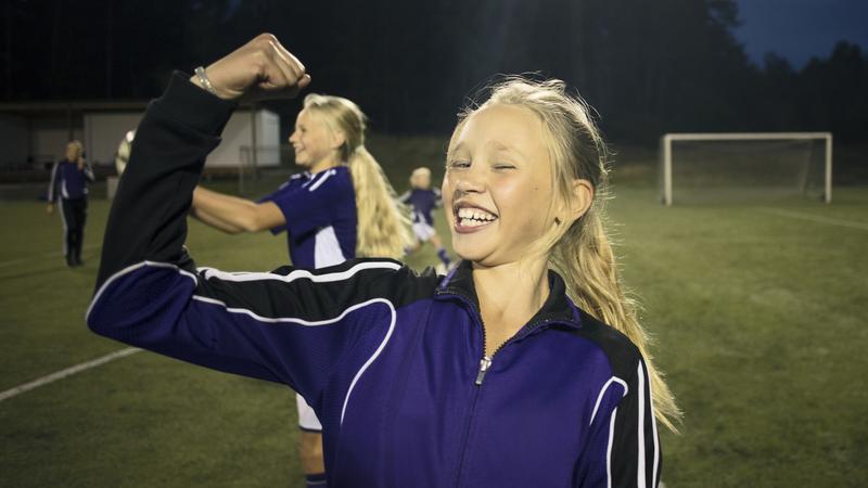 Flicka som ler och spänner musklerna på en fotbollsplan