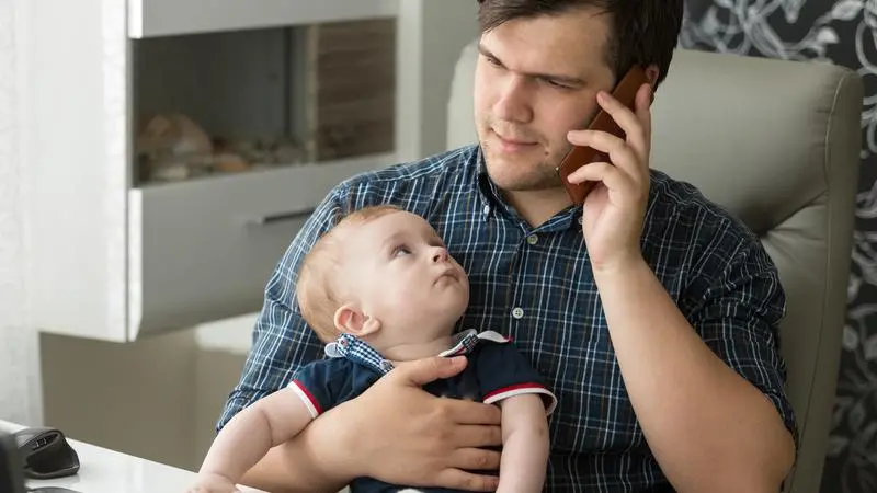 Man som sitter vid ett skrivbord och talar i telefon med en bebis i knät