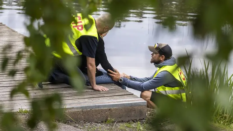 Två personer i gul väst stöttar en person som sitter på en brygga.