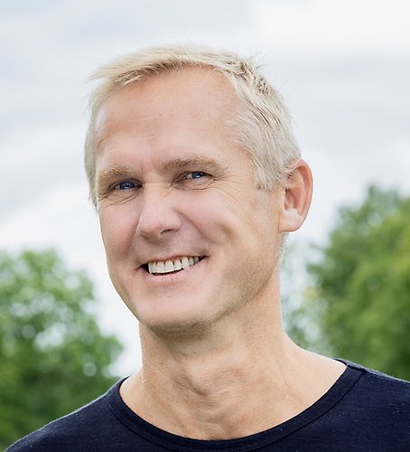 En man i svart skjorta ler mot kameran.