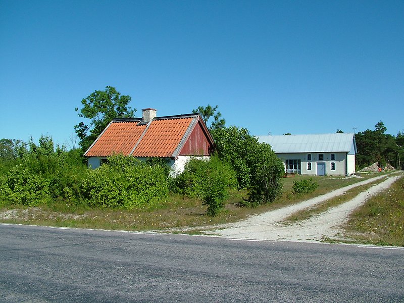 En lite stuga stående vid vägen, längre bort finns det en vit lada med blåa fönster, blåa dörrar och ett plåttak.